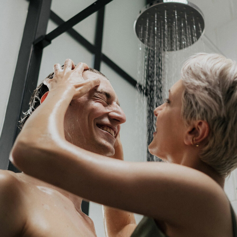 Paar unter einer modernen Regendusche lacht sich an, während sie ihm liebevoll die Haare wäscht – stilvolles, barrierearmes Badezimmer mit Glasduschkabine von Kinedo.