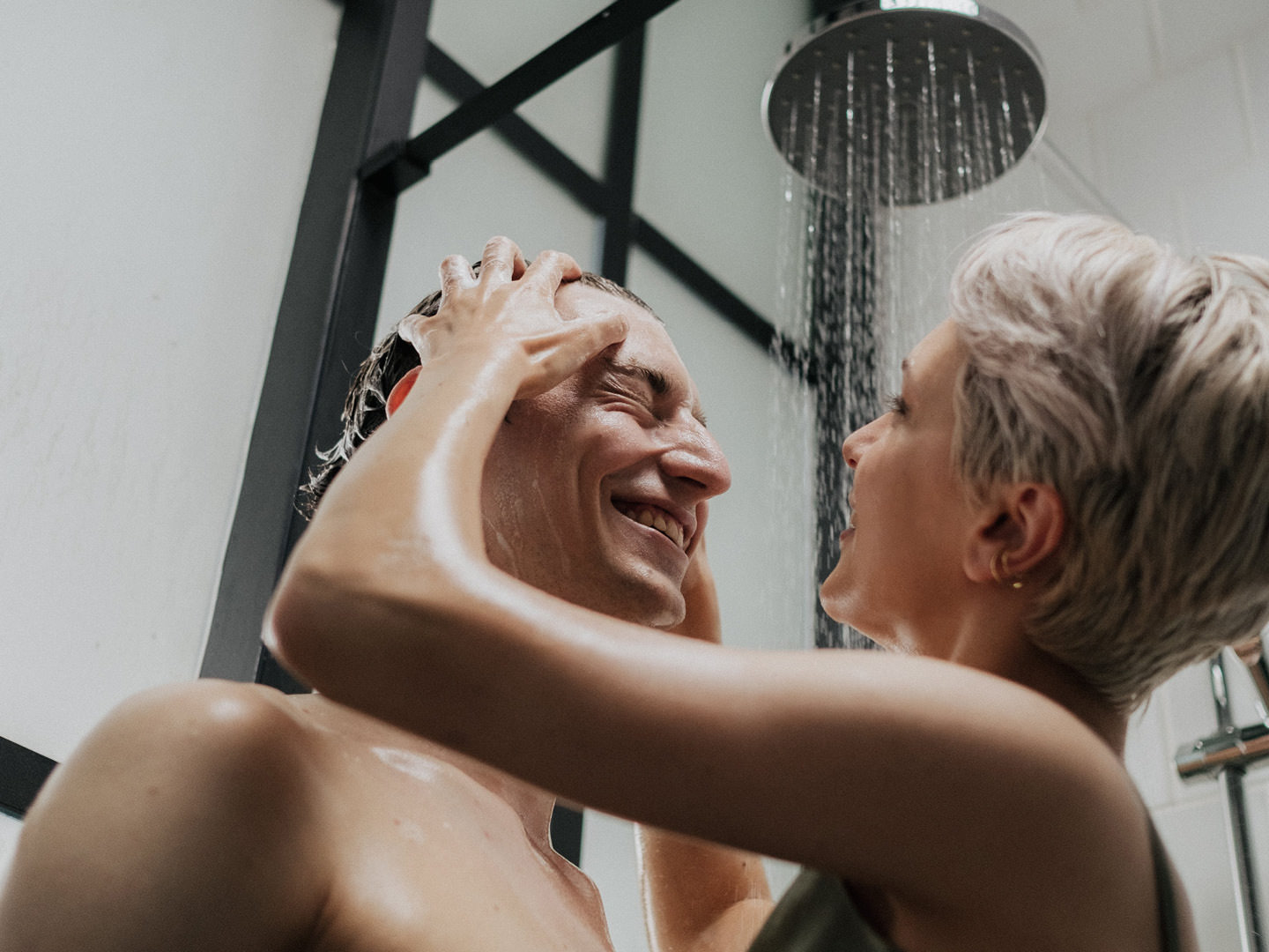 Paar unter einer modernen Regendusche lacht sich an, während sie ihm liebevoll die Haare wäscht – stilvolles, barrierearmes Badezimmer mit Glasduschkabine von Kinedo.